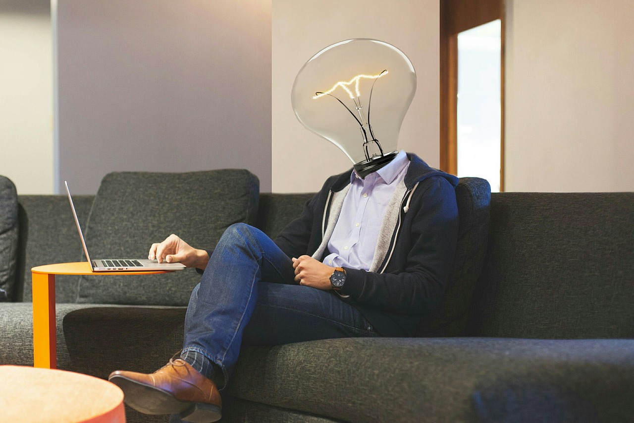 A strange image of a person with a lightbulb for a head sitting on a sofa working out something on a laptop computer. 